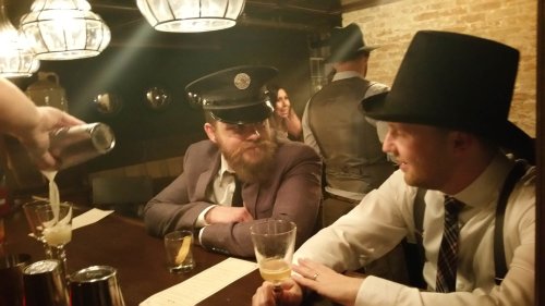 Two men in vintage attire sit at a bar, one wearing a police hat, while a bartender pours a drink.
