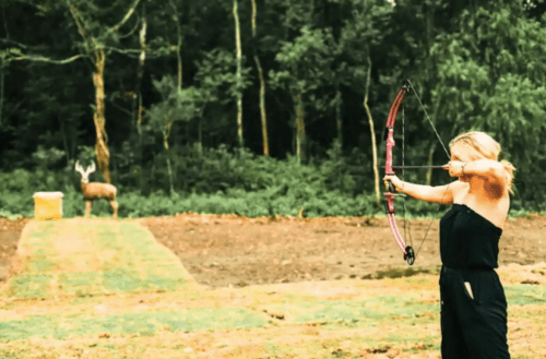A person aiming a bow and arrow at a target in a forested area, with a deer in the background.