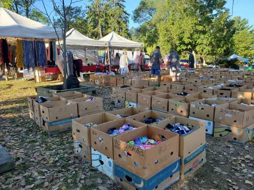 A market scene with cardboard boxes filled with items, surrounded by tents and people browsing among the trees.