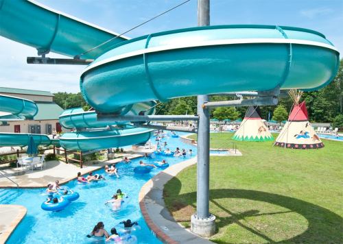 A water park scene featuring blue slides, a lazy river with people floating, and colorful teepees in the background.