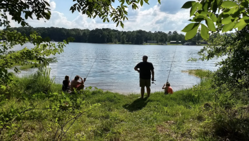 A person stands by a lake while two others fish from the shore, surrounded by greenery and a sunny sky.