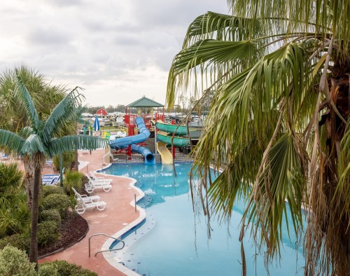A vibrant pool area with water slides, surrounded by palm trees and lounge chairs, under a cloudy sky.