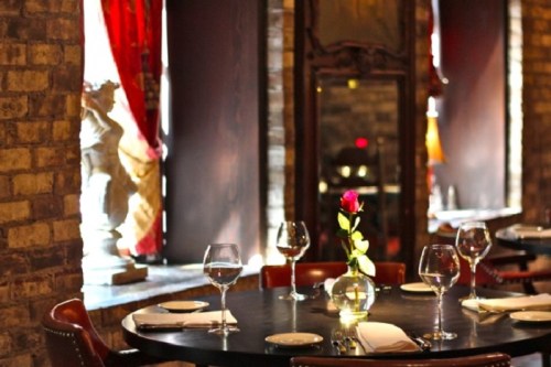 Elegant restaurant table set for dining, featuring a rose centerpiece and wine glasses, with warm lighting and brick walls.