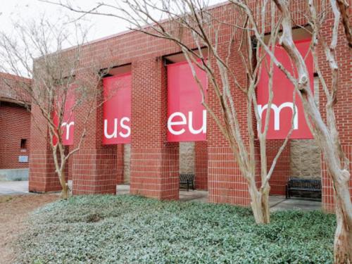 Exterior view of a brick building with large red banners displaying the words "us" and "eu" among trees and greenery.