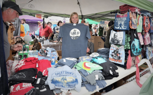 A vendor smiles while holding a "Be Wild" t-shirt at a market booth filled with colorful apparel and accessories.