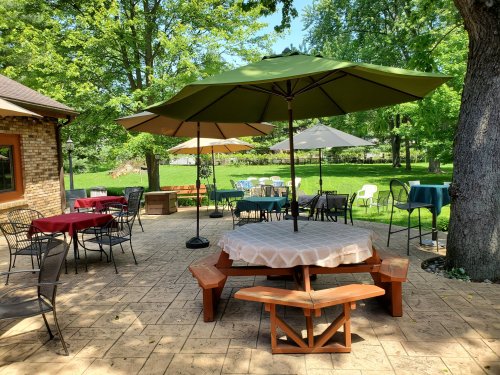 Outdoor dining area with green umbrellas, tables, and chairs surrounded by trees and a grassy lawn.