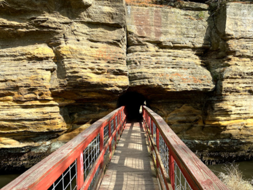 A wooden bridge leads into a dark cave surrounded by layered rock formations.
