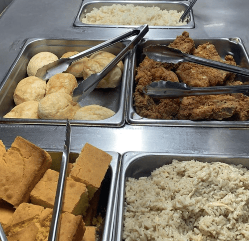 A buffet with trays of fried chicken, rice, cornbread, and rolls, with tongs for serving.