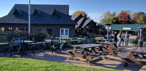 Outdoor seating area with picnic tables and chairs, surrounded by buildings and trees in a sunny setting.
