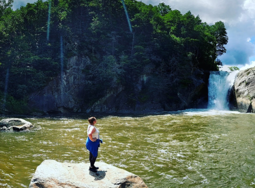 A person stands on a rock by a river, gazing at a waterfall surrounded by lush greenery and a cloudy sky.