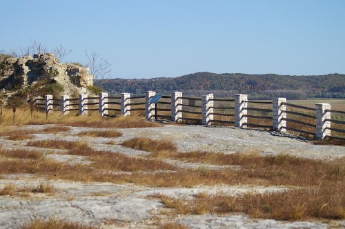 A rocky landscape with a wooden fence and distant hills under a clear blue sky. Grass grows in the foreground.