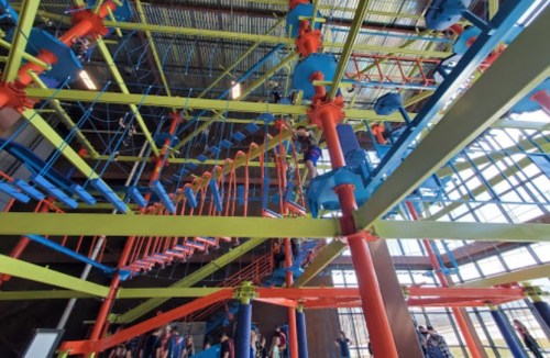 A colorful indoor climbing structure with bright blue, green, and orange elements, filled with people navigating the course.