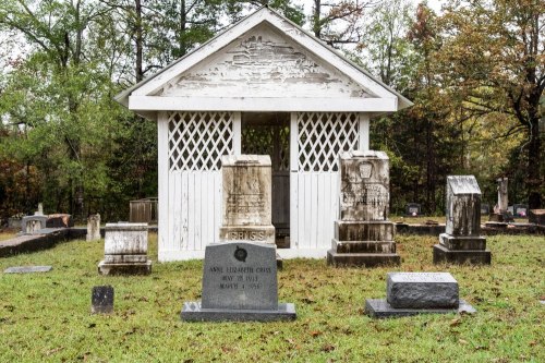 A small white mausoleum surrounded by gravestones in a wooded cemetery, with autumn foliage in the background.