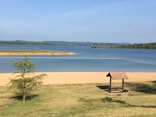 A serene lakeside view with a sandy beach, a small shelter, and distant hills under a clear blue sky.