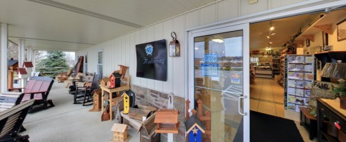 A welcoming storefront with wooden crafts, birdhouses, and seating on a porch. The entrance features a "Welcome" sign.