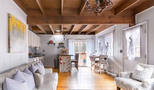 Cozy living room with wooden beams, light-colored furniture, and a view of a deck through large windows.