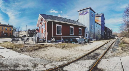 A red building with a porch next to a blue mill, alongside a gravel path and railway tracks under a clear blue sky.