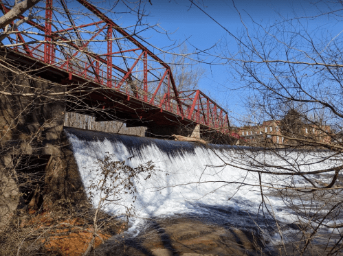 A red metal bridge spans a waterfall, surrounded by trees and a clear blue sky. Water cascades over the dam below.