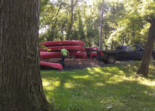 A person in a green shirt is near stacked red canoes by a black truck in a wooded area.