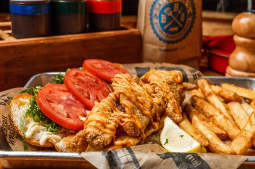 A plate featuring a sandwich with lettuce, tomatoes, and fried fish, accompanied by crispy fries and a lemon wedge.