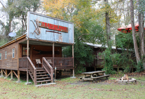 A rustic wooden cabin with a "Trading Post" sign, surrounded by trees and a picnic table in front.