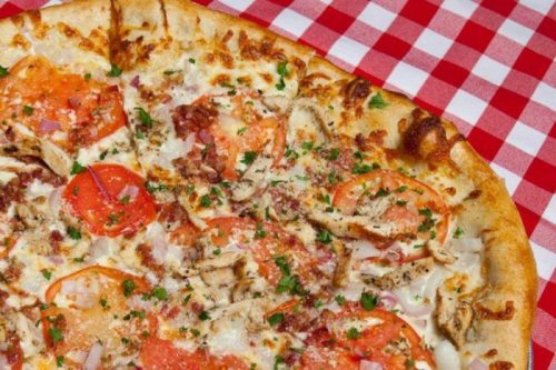 Close-up of a pizza topped with tomatoes, chicken, cheese, and herbs on a red and white checkered tablecloth.