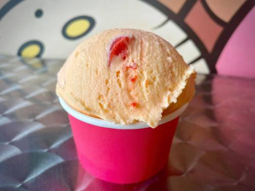 A scoop of peach ice cream with bits of fruit in a pink cup, set against a colorful background.