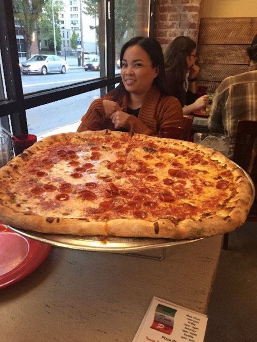 A woman sits at a table with a large pepperoni pizza on a tray in front of her, smiling in a cozy restaurant setting.