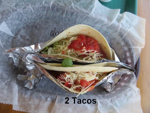 Two tacos filled with shredded cheese, lettuce, and salsa, wrapped in foil and placed on a paper-lined tray.