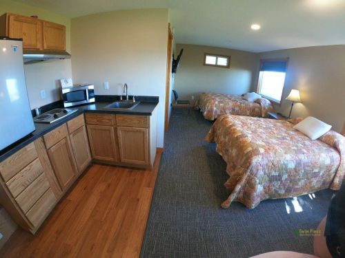 Cozy hotel room with two beds, kitchenette, and a window, featuring warm wood tones and a simple layout.