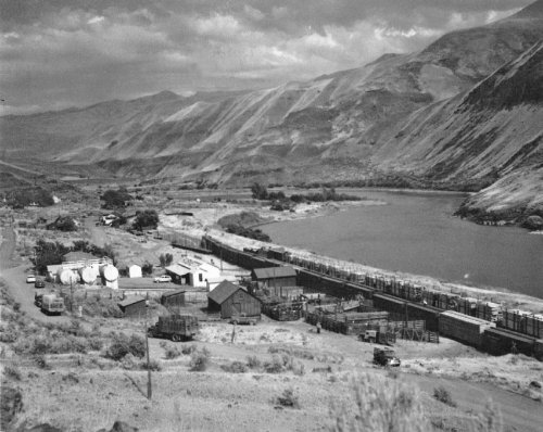 Black and white landscape of a river valley with buildings, tanks, and mountains in the background.