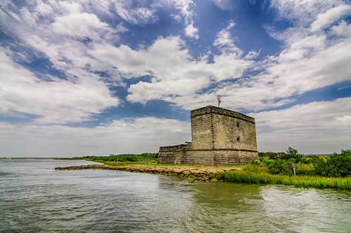 A historic stone fort stands by a river under a cloudy sky, surrounded by greenery and water.