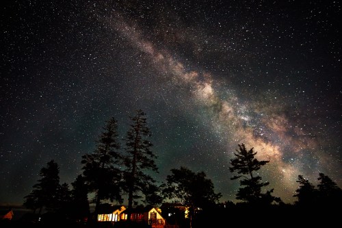 A starry night sky with the Milky Way above silhouetted trees and a warmly lit house.