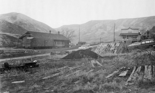Black and white photo of a rural landscape with buildings, mountains, and scattered construction materials.