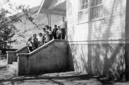 A group of children stands on the steps of a house, with trees and hills in the background, in a vintage black-and-white photo.