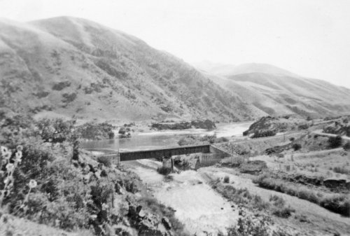 Black and white landscape featuring a river, a bridge, and rolling hills in the background.