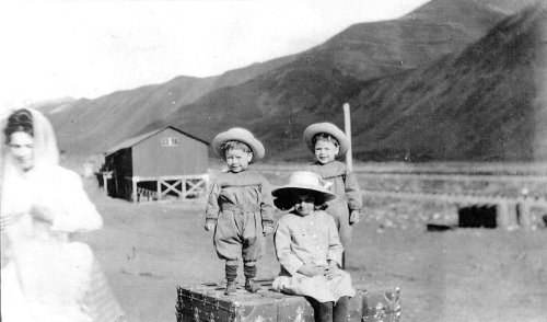 Three children in hats stand and sit on a crate outdoors, with a woman in a white dress walking nearby and mountains in the background.