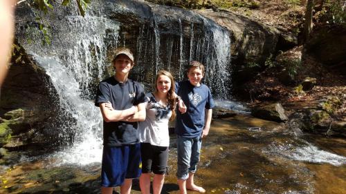 Three young people stand by a waterfall in a forest, smiling and posing for the photo. Sunlight filters through the trees.