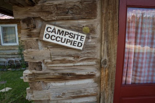 A wooden cabin wall with a sign that reads "Campsite Occupied" hanging near a red door and a window with checkered curtains.