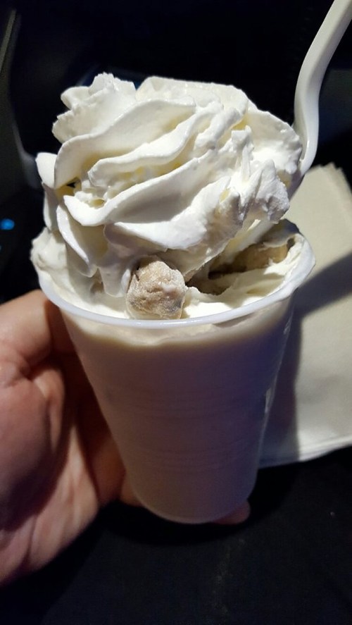 A hand holding a cup of creamy dessert topped with whipped cream and chunks of cookie.