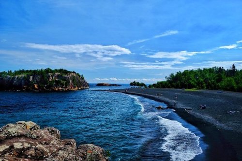 A scenic view of a beach with a rocky shoreline, clear blue water, and lush green trees under a bright sky.