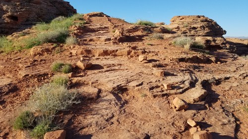Rocky terrain with scattered vegetation and a clear blue sky, leading up a slope.