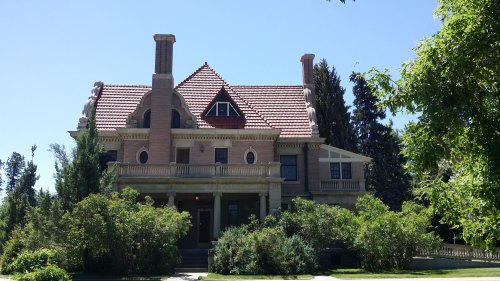 A large, historic house with a red tile roof, surrounded by greenery and tall trees under a clear blue sky.