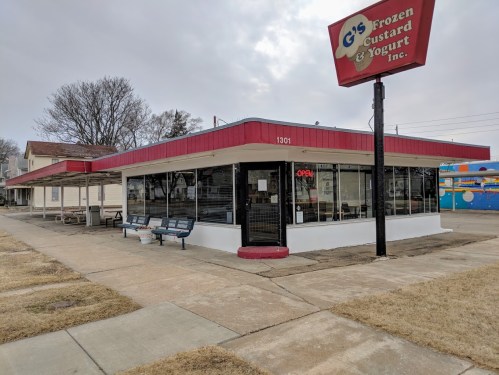 Exterior of G's Frozen Custard & Yogurt, featuring a red awning and "Open" sign, with benches outside.