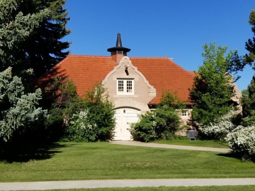 A charming stone house with a red tile roof, surrounded by greenery and trees under a clear blue sky.