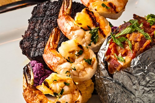 Grilled shrimp and steak served with colorful vegetables and a baked potato wrapped in foil.