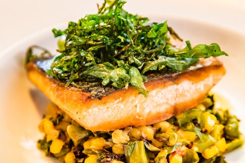 A piece of grilled fish topped with crispy greens, served on a bed of corn and vegetables.