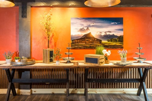 A rustic table display with plants and decor against a vibrant orange wall featuring a mountain landscape painting.