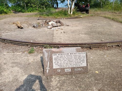 A stone plaque at the site of the Keweenaw Rocket Range, with remnants of a launch pad and surrounding nature.