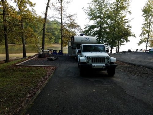 A white Jeep parked next to a camper by a serene lake, surrounded by trees in autumn colors.